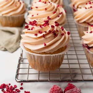 Cupcakes with raspberries decorated with raspberry cream cheese frosting are standing on a wire rack.