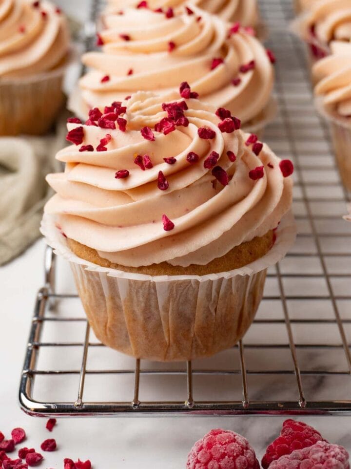 Cupcakes with raspberries decorated with raspberry cream cheese frosting are standing on a wire rack.