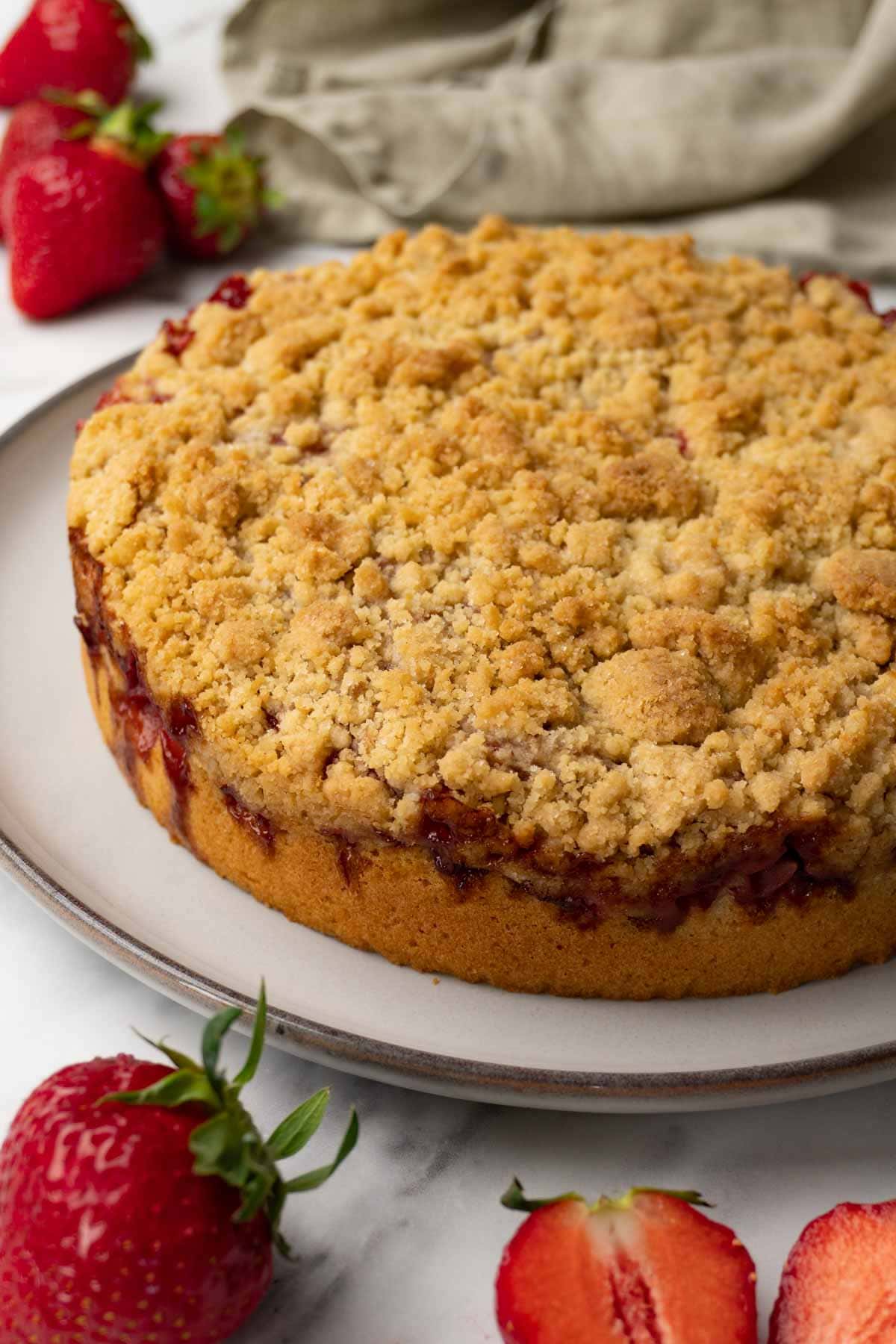 A freshly baked strawberry cake with crumble topping on a round plate; fresh strawberries are lying around the plate.