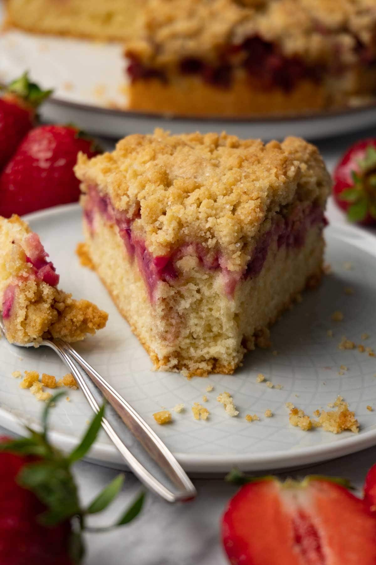 A piece of strawberry cake with crumble topping on a plate; one bite was taken with a silver fork. Fresh strawberries are lying around the plate.