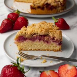 A piece of strawberry cake with crumble topping on a round plate; fresh strawberries are lying around the plate.