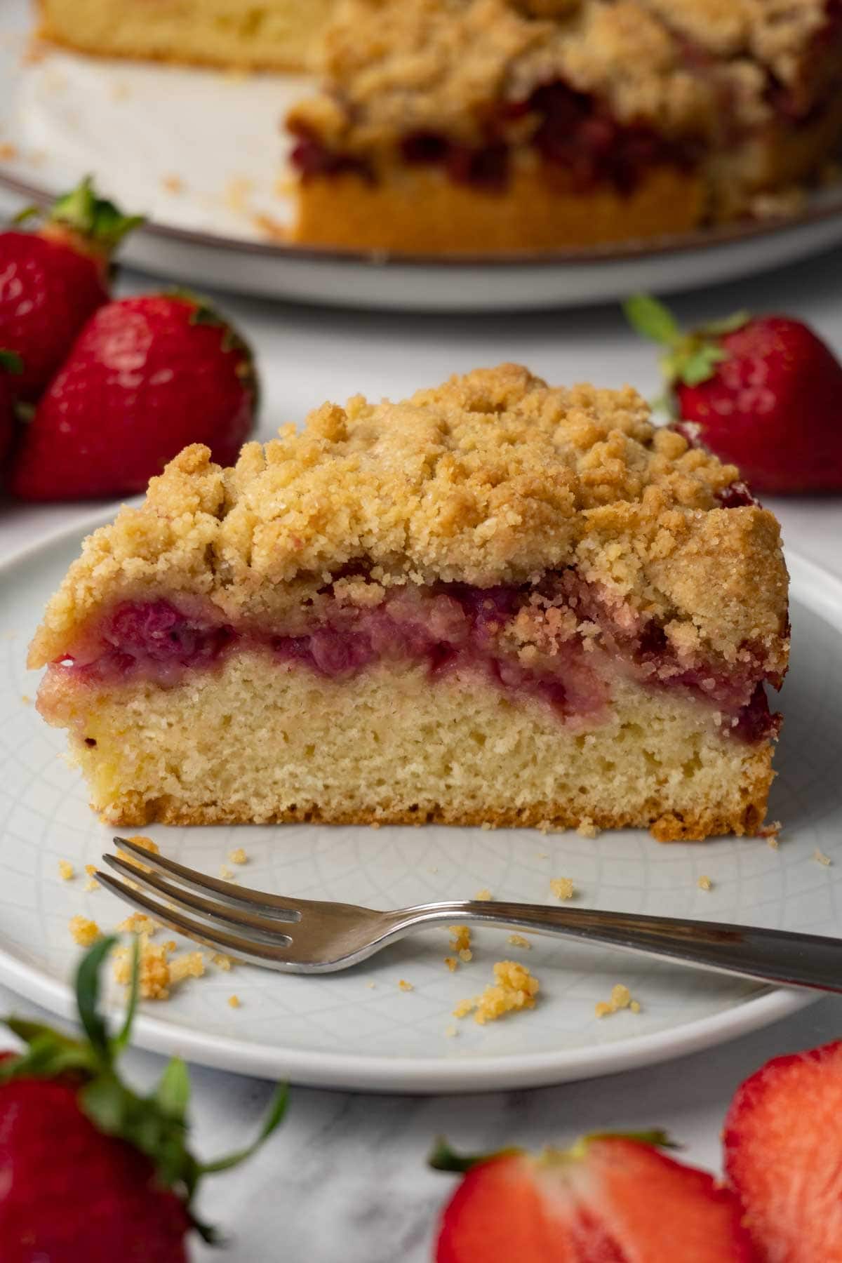 A piece of strawberry cake with crumble topping on a round plate; fresh strawberries are lying around the plate.