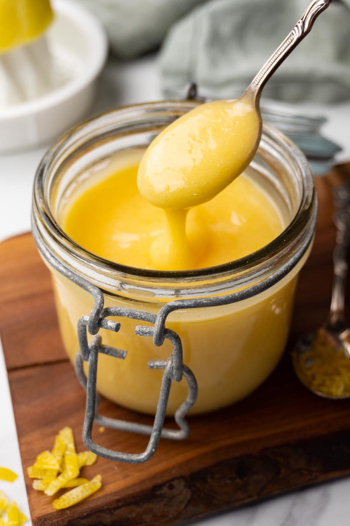 A glass jar filled with lemon curd is standing on a wooden board. A silver spoon is taking out some of the curd.