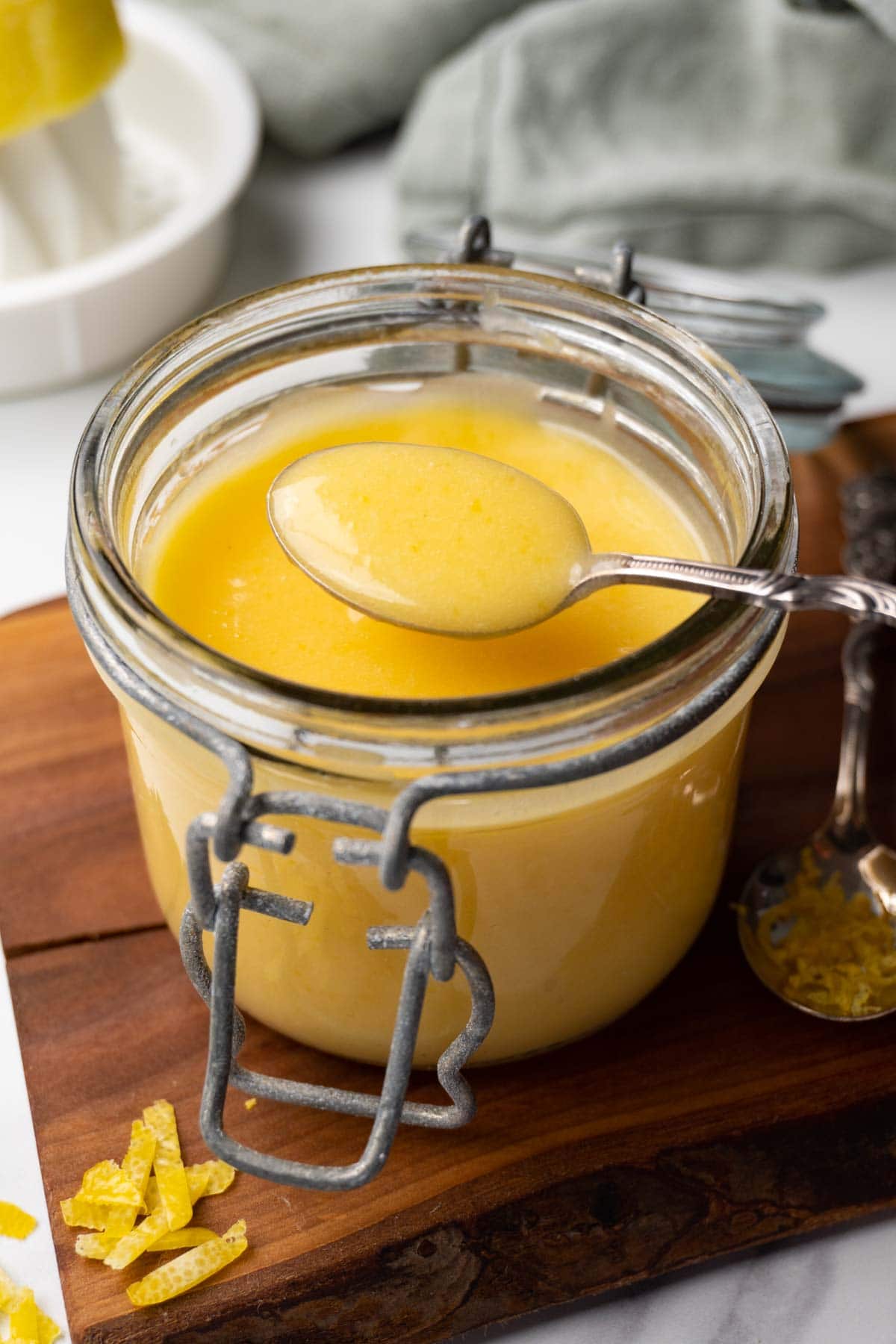 A glass jar filled with lemon curd is standing on a wooden board. A silver spoon with some curd is hovering over the jar.