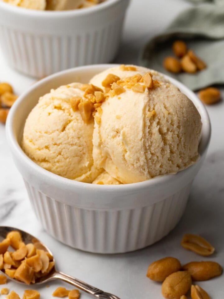 Ice cream topped with crushed peanuts in a white ramekin. Roasted peanuts are lying around.