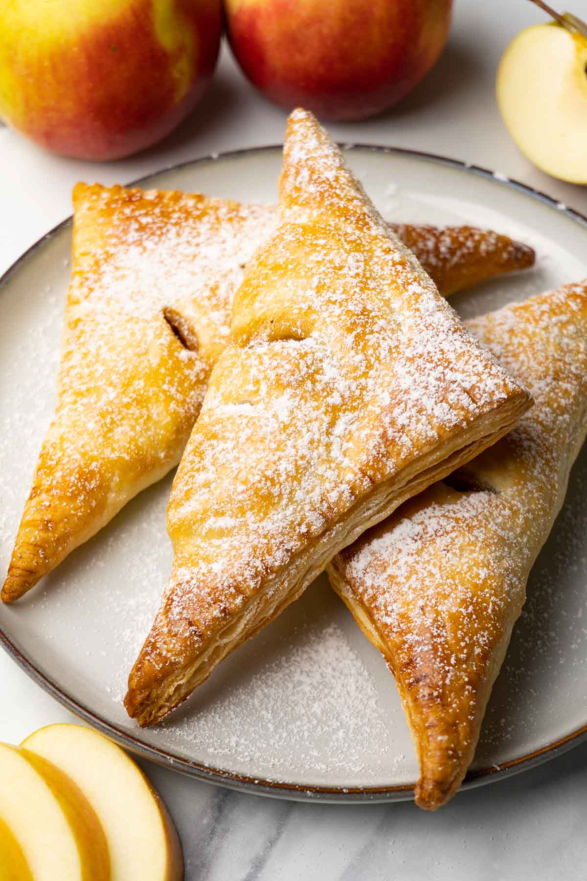 Apple turnovers dusted in powdered sugar on a plate. Fresh apples are lying around.