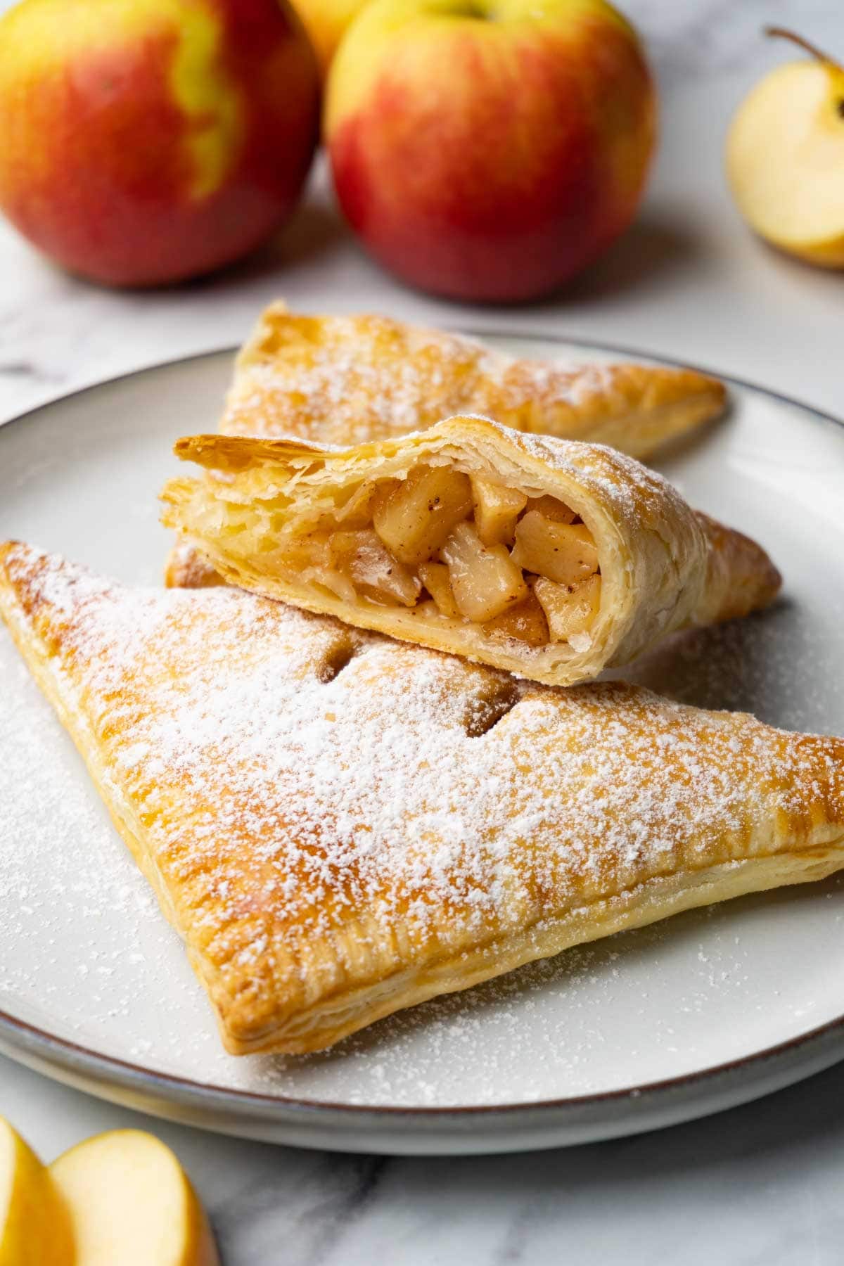 Apple turnovers dusted in powdered sugar on a plate. One turnover is cut in half. Fresh apples are lying around.