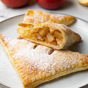 Apple turnovers dusted in powdered sugar on a plate. One turnover is cut in half.