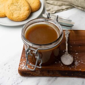 A glass jar filled with butterscotch sauce on a wooden board with silver spoon on it.