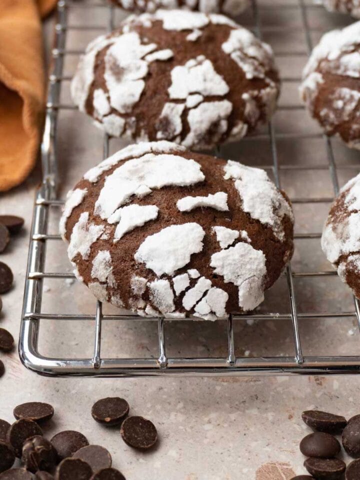 Chocolate cookies covered in powdered sugar are on a cooling rack. Chocolate chips are lying around.