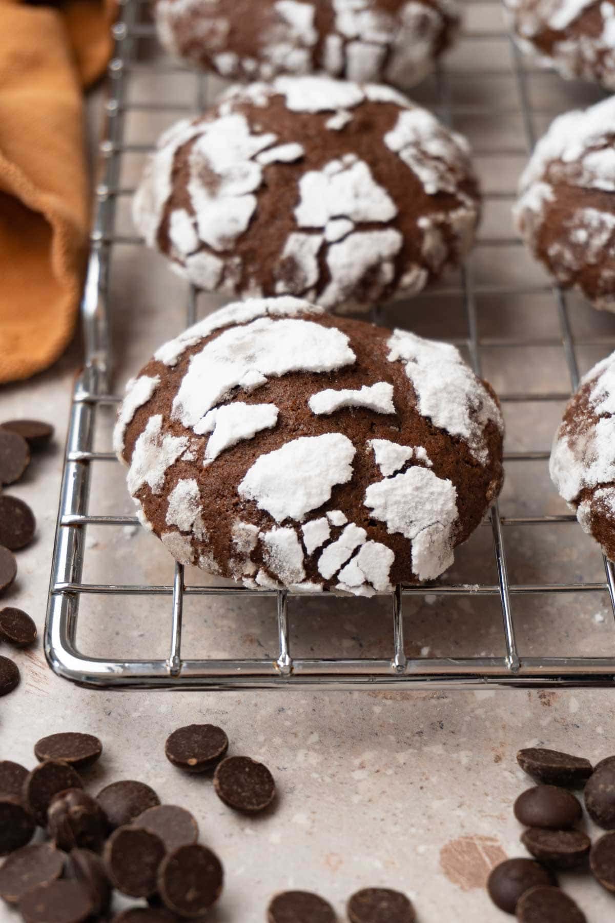 Chocolate cookies covered in powdered sugar are on a cooling rack. Chocolate chips are lying around.