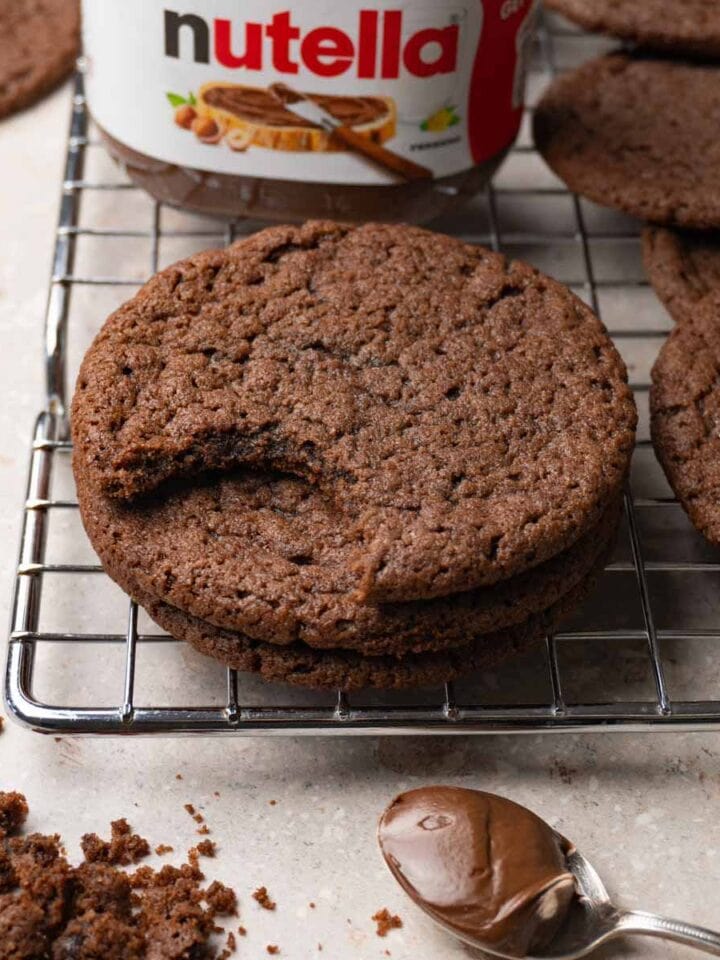 Round chocolate cookies on a metal cooling rack; one bite was taken from one of the cookies. A jar of Nutella is standing on the background.