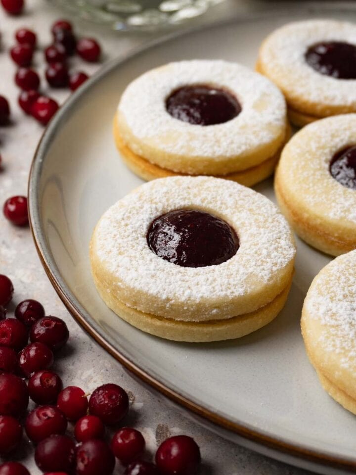 Round sandwich cookies with cranberry jam and dusted with powdered sugar are served on a round plate. Fresh cranberries are lying around.