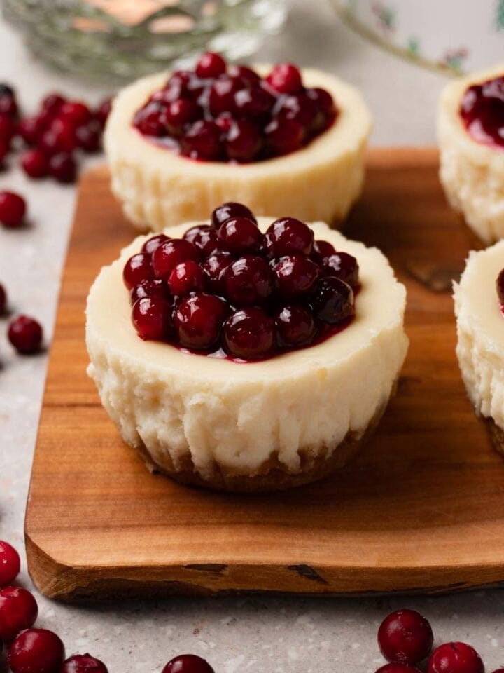 Mini cheesecakes with cranberry jam topping on a wooden serving board. Fresh cranberries are lying around the board.