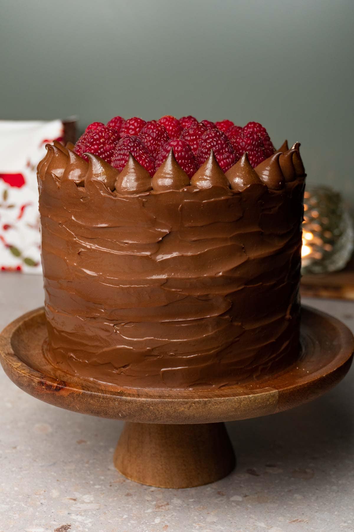 A rustic-looking chocolate cake on a wooden plate covered with silky chocolate cream and decorated with fresh raspberries.