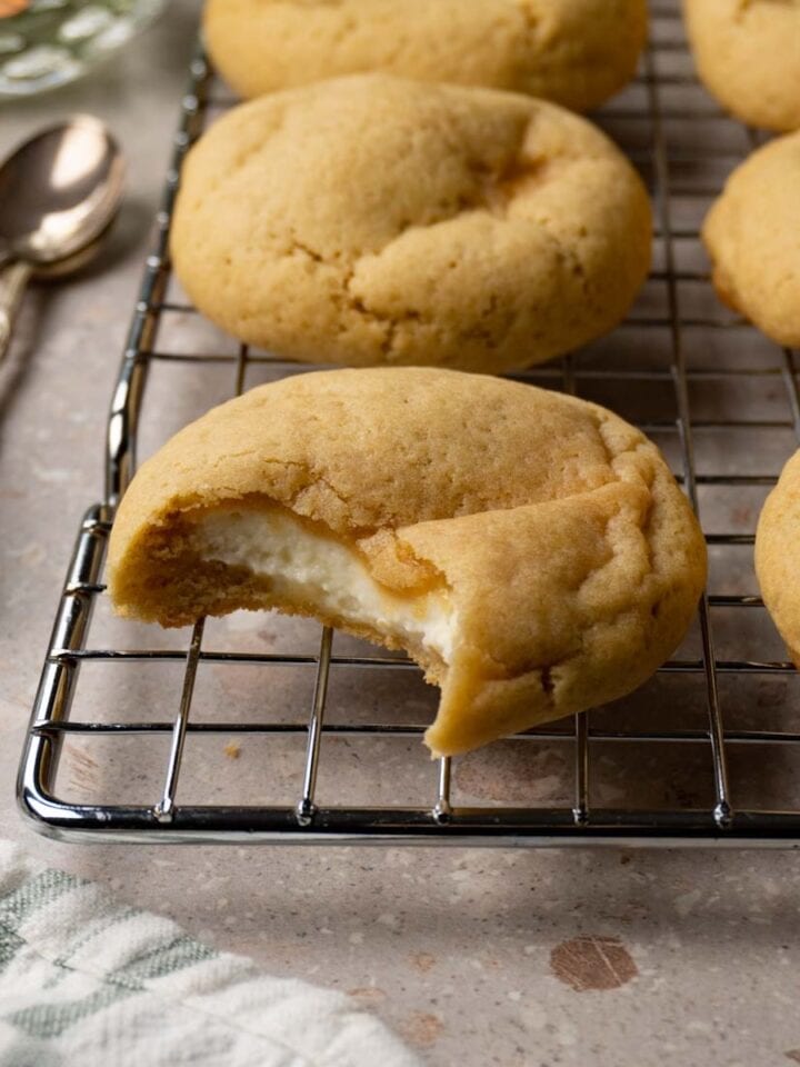 Vanilla cookies with cheesecake filling on a metal cooling rack. One bite was taken from one of the cookies.