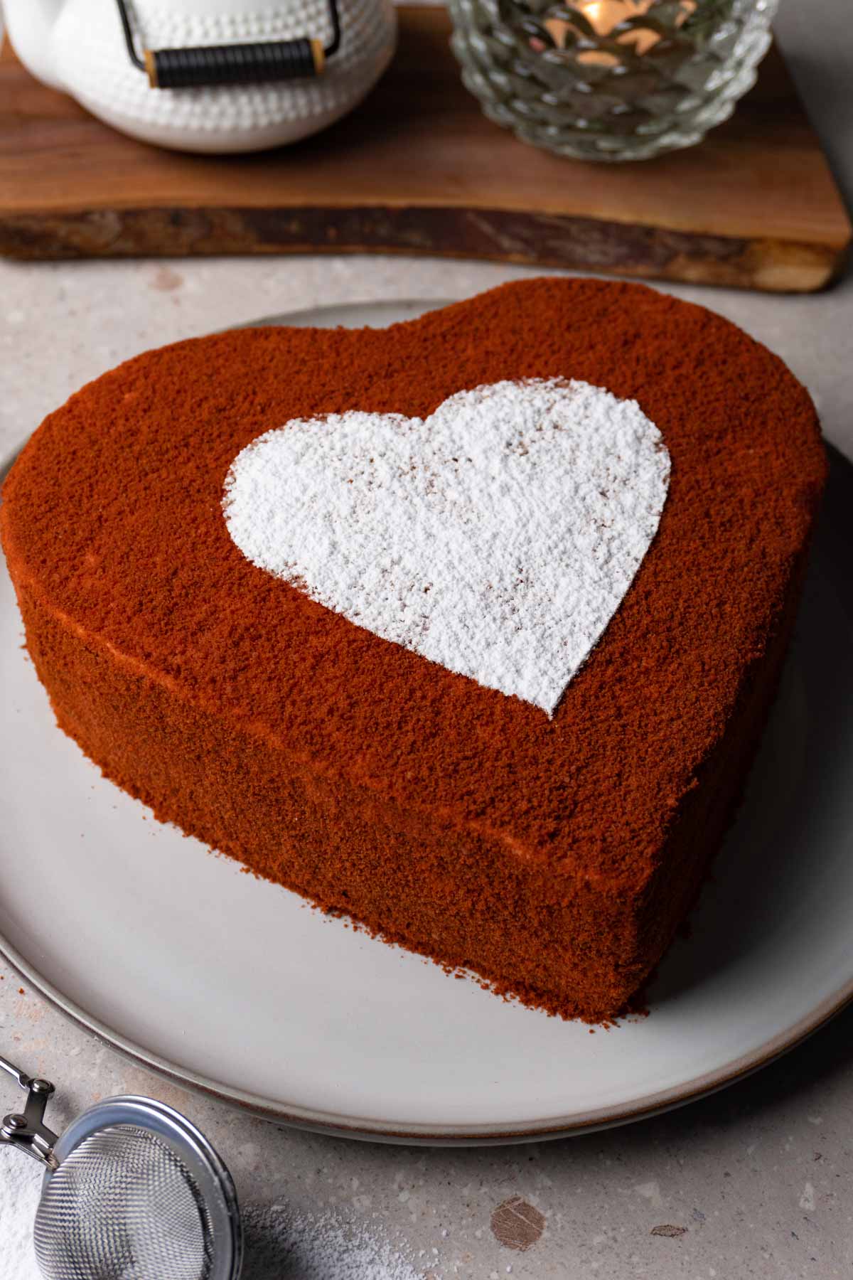 Red velvet heart shaped cake decorated with a white powder sugar heart on top on a large round plate..