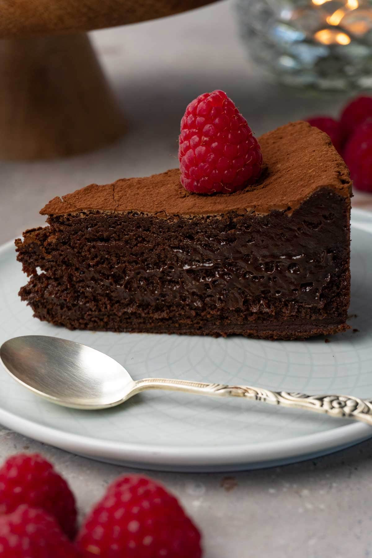 A piece of chocolate cake dusted with cocoa powder and topped with fresh raspberries on a plate with a silver spoon.