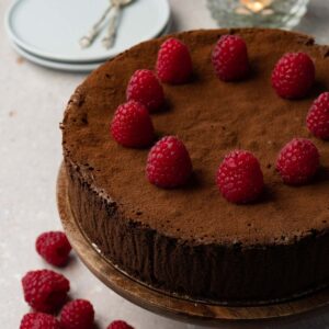 A chocolate cake dusted with cocoa powder and topped with fresh raspberries on a wooden cake stand.