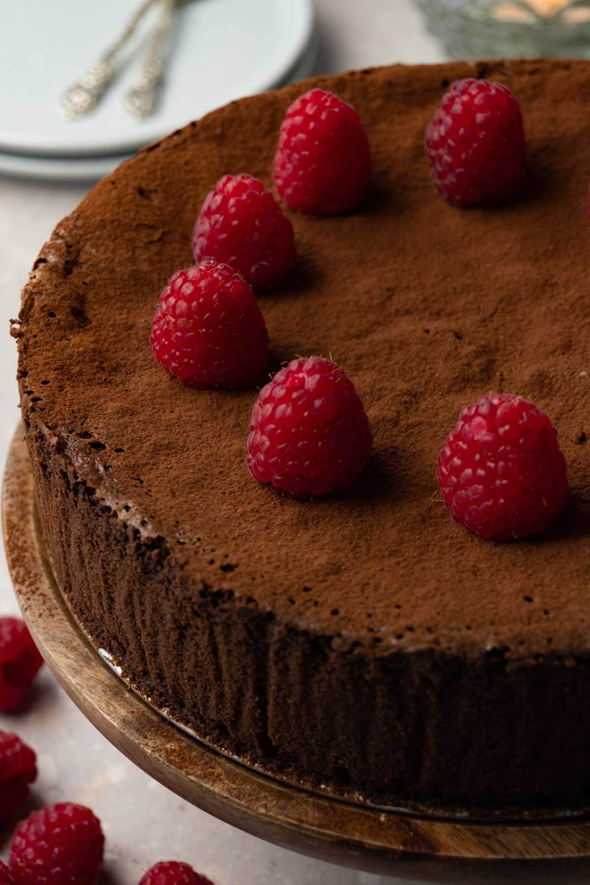 A chocolate cake dusted with cocoa powder and topped with fresh raspberries on a wooden cake stand.