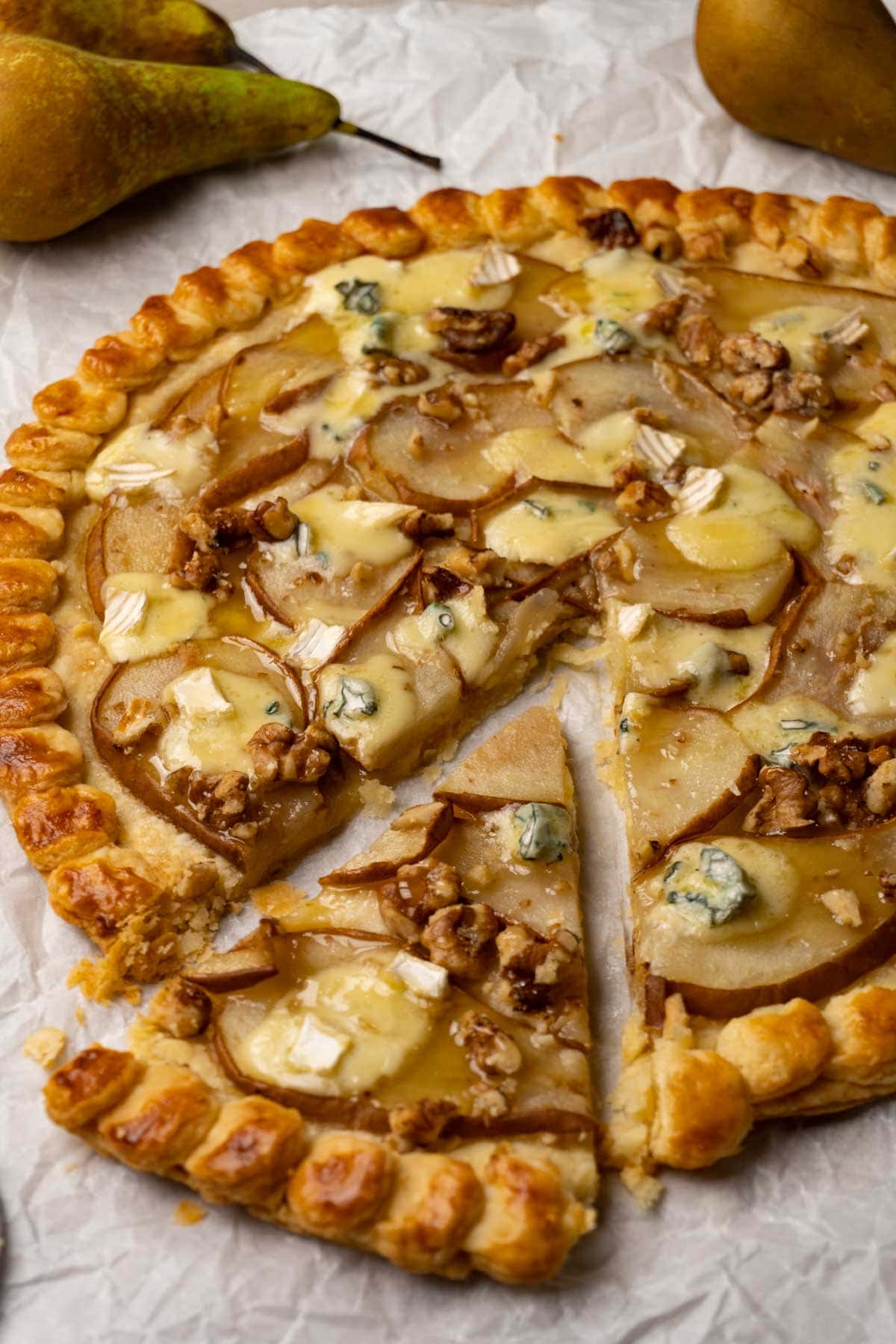 A round puff pastry galette with pears, walnuts, blue cheese and honey on a white parchment paper. One slice was taken.