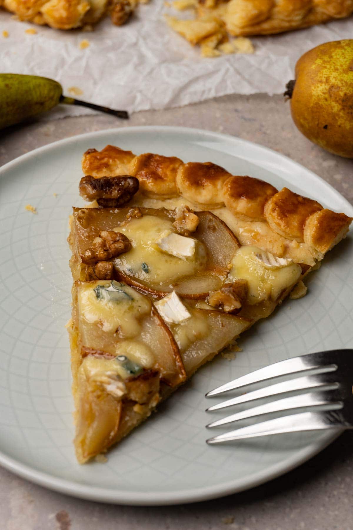 A slice of galette with pears, walnuts, blue cheese and honey served on a small round plate with silver fork.