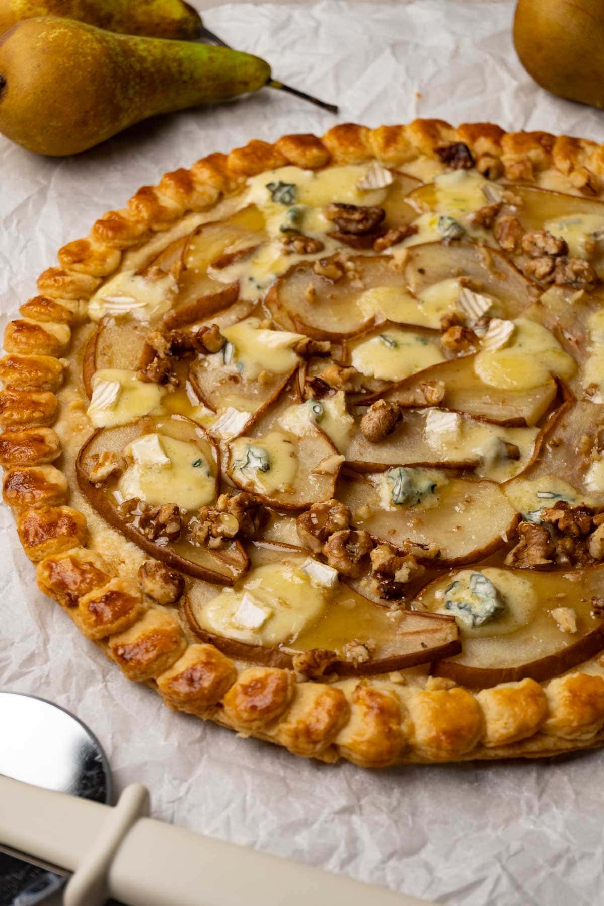 A round puff pastry galette with pears, walnuts, blue cheese and honey on a white parchment paper. Fresh pears and a pizza cutter are lying next to the galette.