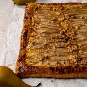 A tart made with puff pastry, pears, walnuts and topped with honey on a white parchment paper.