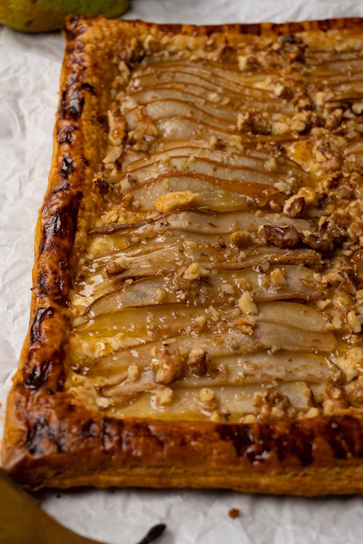 A tart made with puff pastry, pears, walnuts and topped with honey on a white parchment paper.