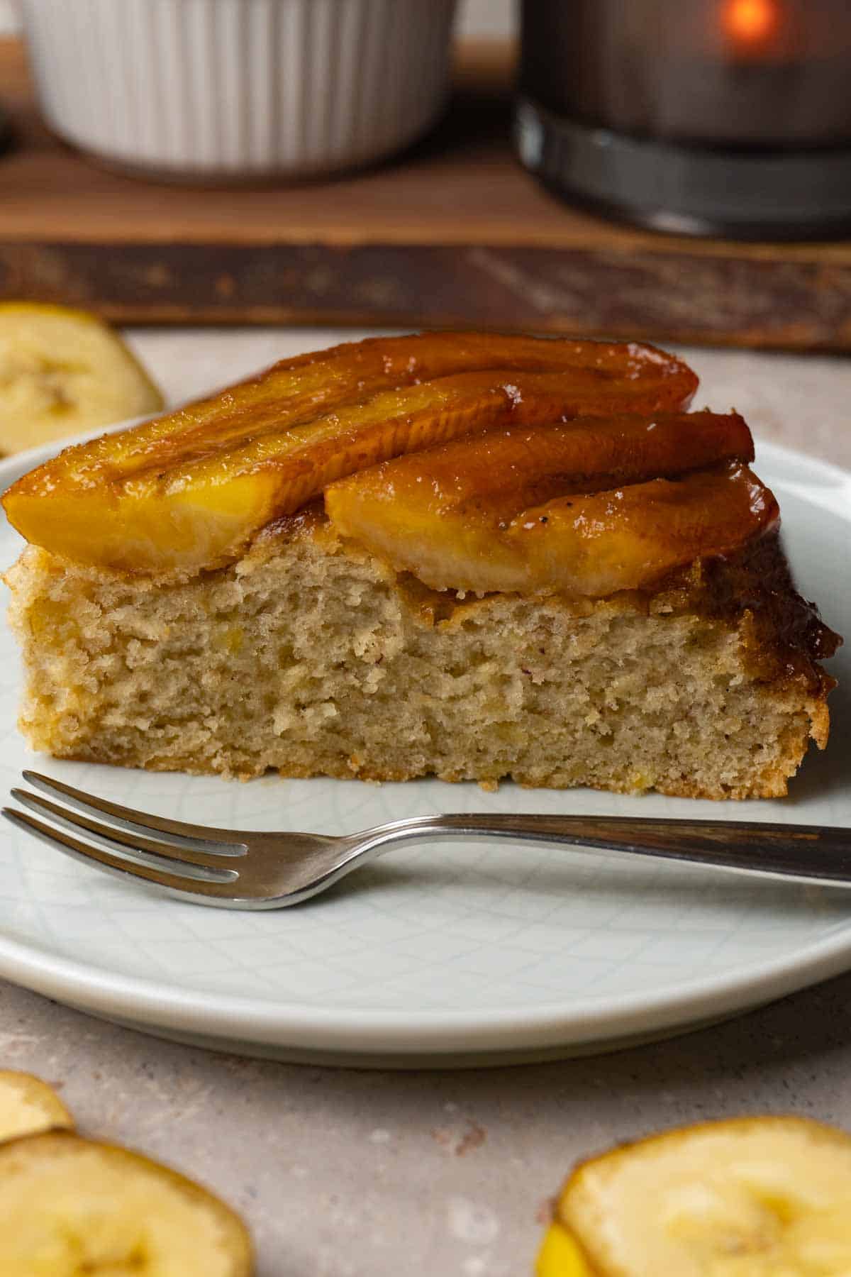 A piece of cake with a caramelized banana topping is served on a small round plate with a silver dessert fork. Slices of fresh banana are lying on the table nearby.