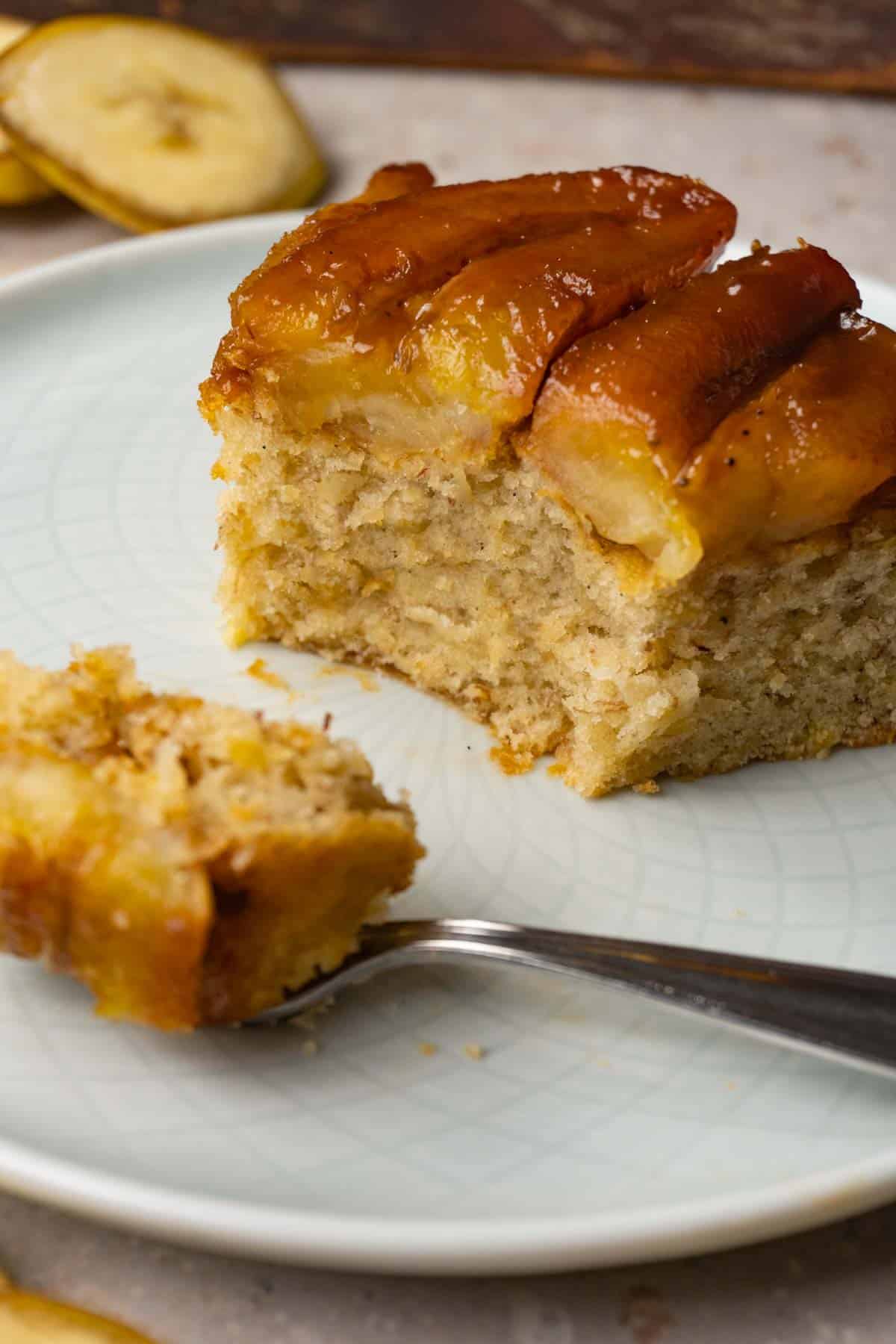 A piece of cake with a caramelized banana topping is served on a small round plate. One bite has been taken with a silver dessert fork.