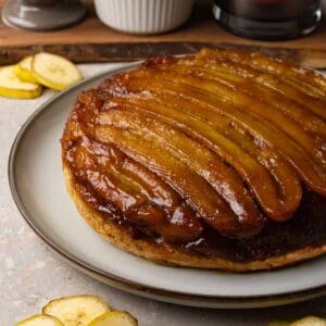 A cake with a caramelized banana topping is served on a large round plate. Slices of fresh banana are lying on the table nearby.