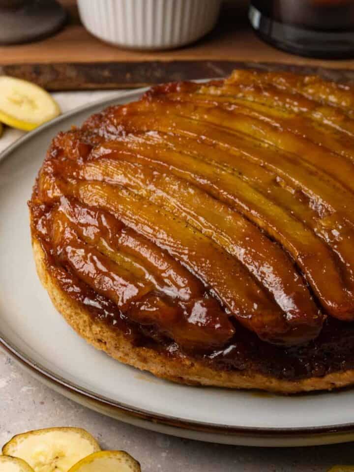 A cake with a caramelized banana topping is served on a large round plate. Slices of fresh banana are lying on the table nearby.