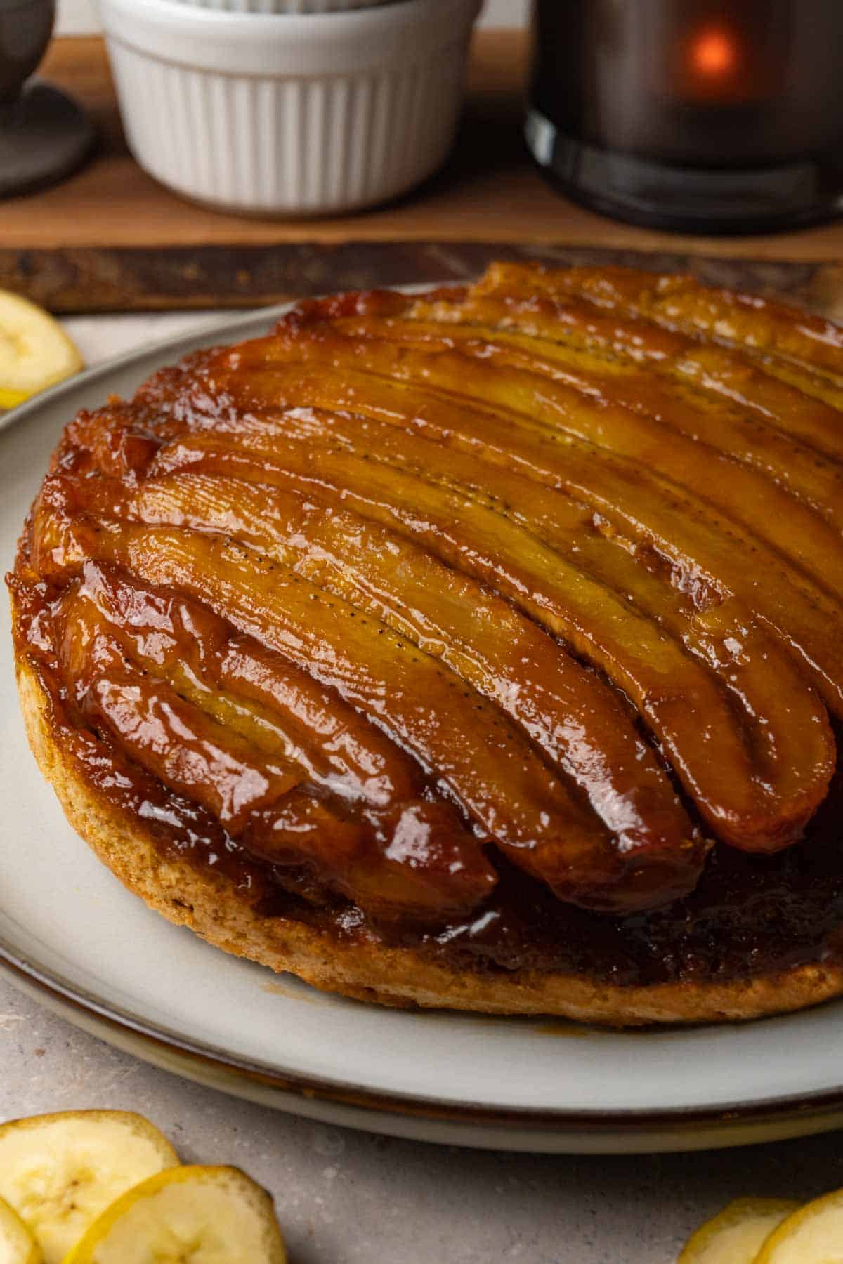 A cake with a caramelized banana topping is served on a large round plate. Slices of fresh banana are lying on the table nearby.
