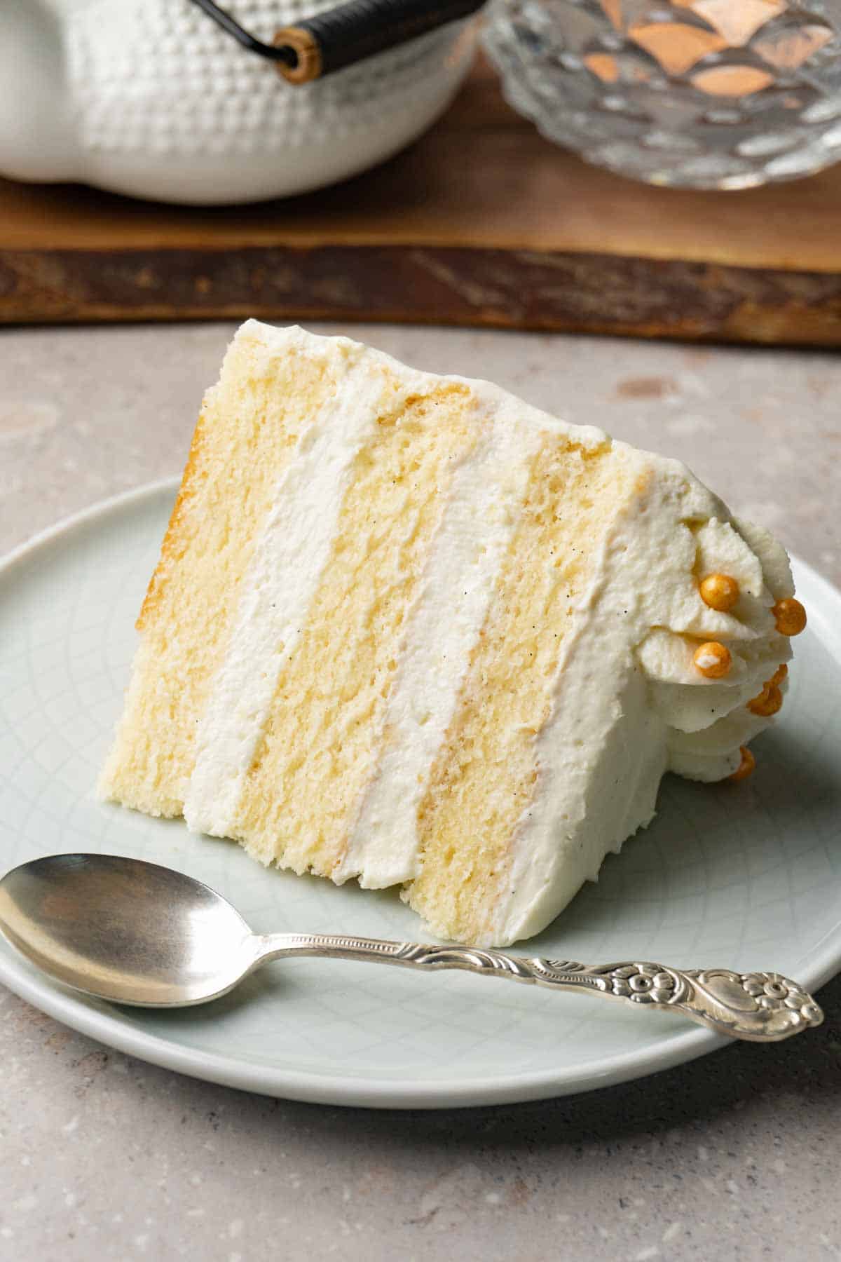 A piece of cake with white frosting sits on a small round plate with a silver spoon.