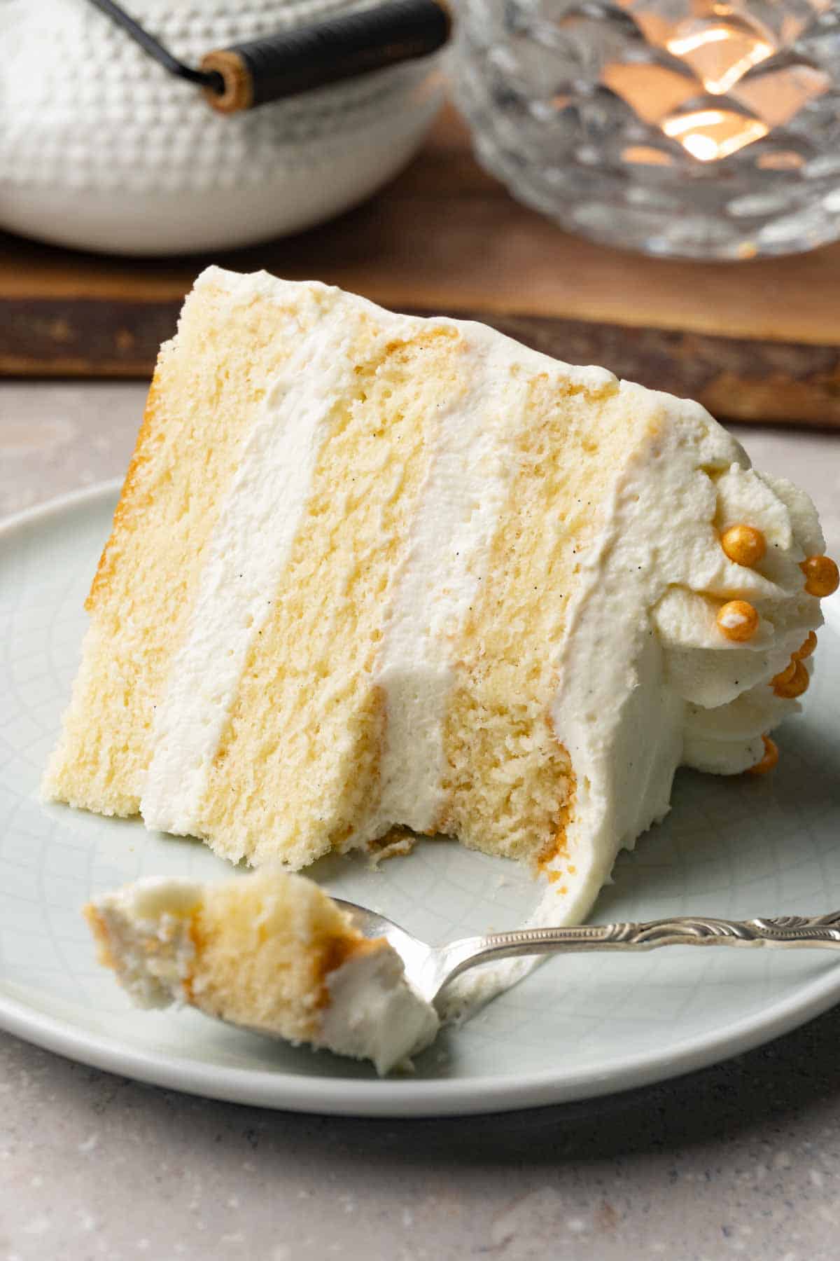A piece of cake with white frosting sits on a small round plate. One bite has been taken with a silver spoon.