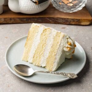 A piece of cake with white frosting sits on a small round plate with a silver spoon.