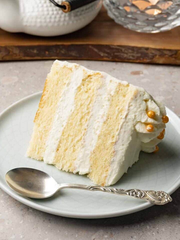 A piece of cake with white frosting sits on a small round plate with a silver spoon.