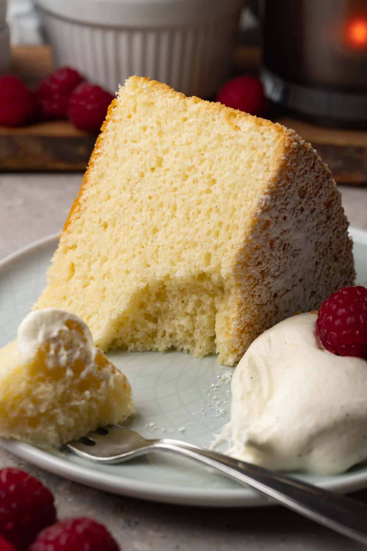 A piece of chiffon cake on a small round plate served with whipped cream and fresh raspberries. One bite has been taken with a silver spoon.