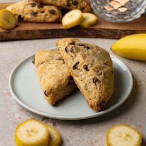 Banana scones with chocolate chips on a small round plate. Slices of fresh bananas are lying around the plate.