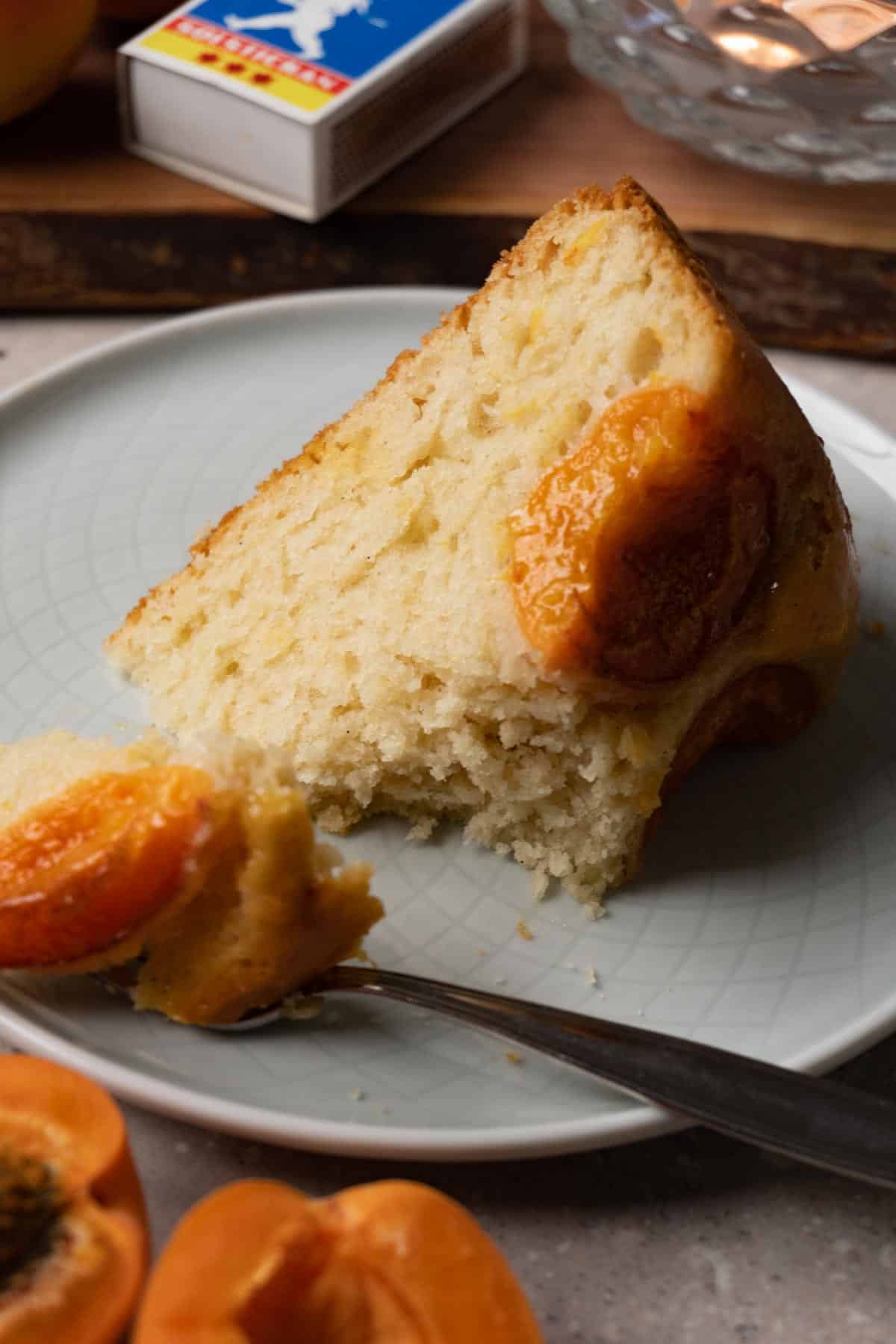 A moist slice of peach cake on a small round plate with a silver dessert fork. One bite has been taken.
