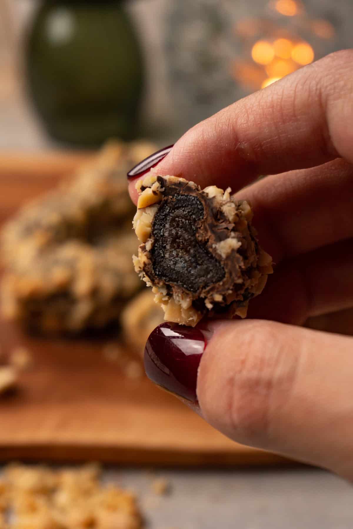 A hand holds a chocolate-covered date, coated in crushed roasted peanuts, cut in half.