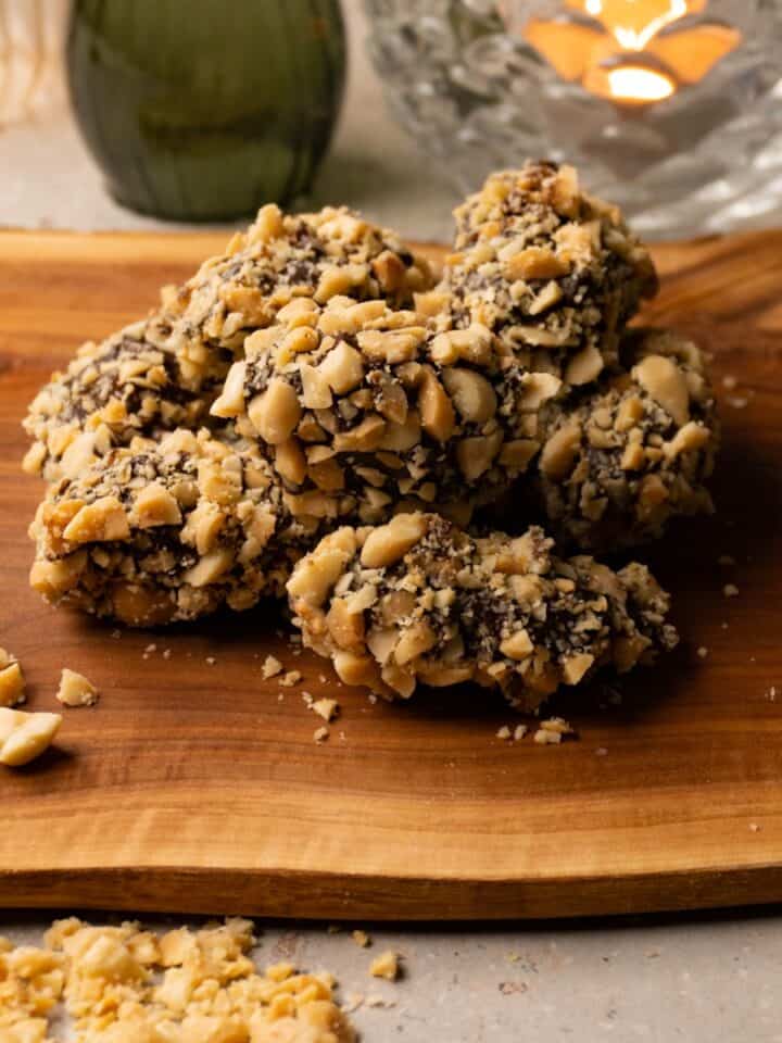 Chocolate-covered dates coated in crushed roasted peanuts are arranged on a wooden board.