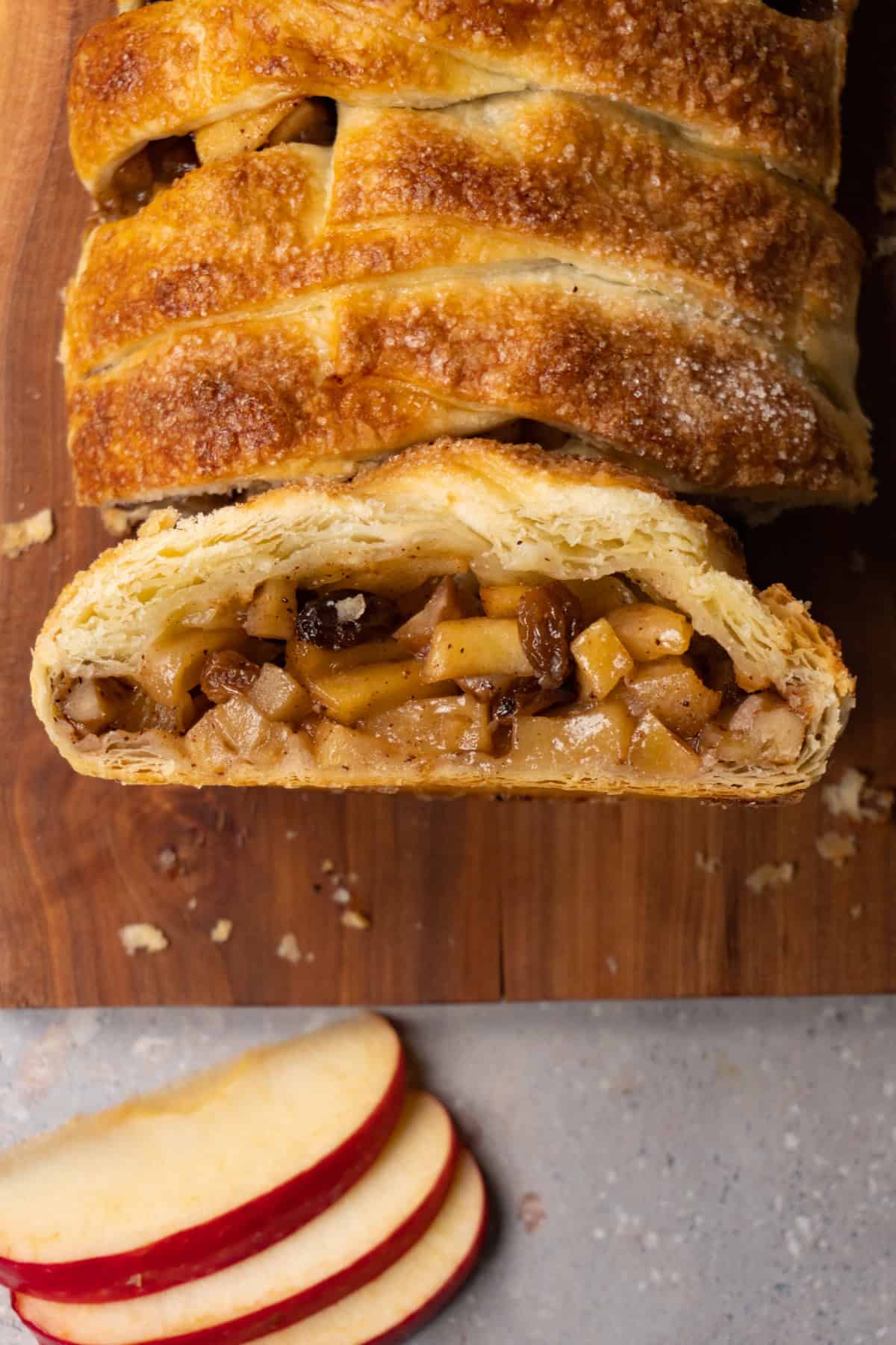 A puff pastry pie in a log shape with a braided pattern is served on a wooden board. One piece has been cut and placed with the cut side facing up.