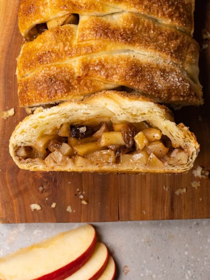 A puff pastry pie in a log shape with a braided pattern is served on a wooden board. One piece has been cut and placed with the cut side facing up.