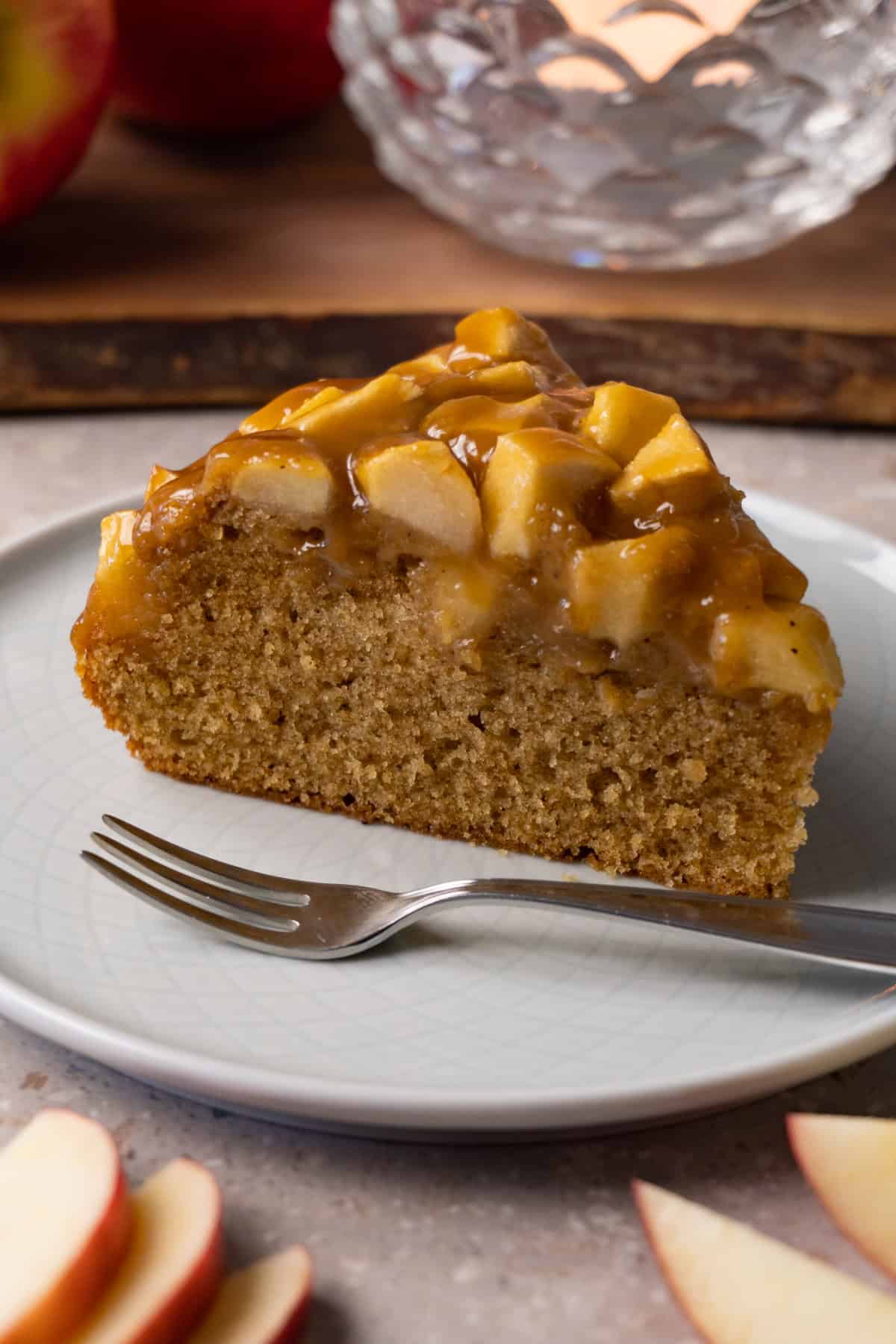 A piece of apple cake topped with toffee sauce sits on a small round plate with a silver dessert fork.
