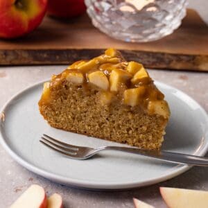 A piece of apple cake topped with toffee sauce sits on a small round plate with a silver dessert fork.
