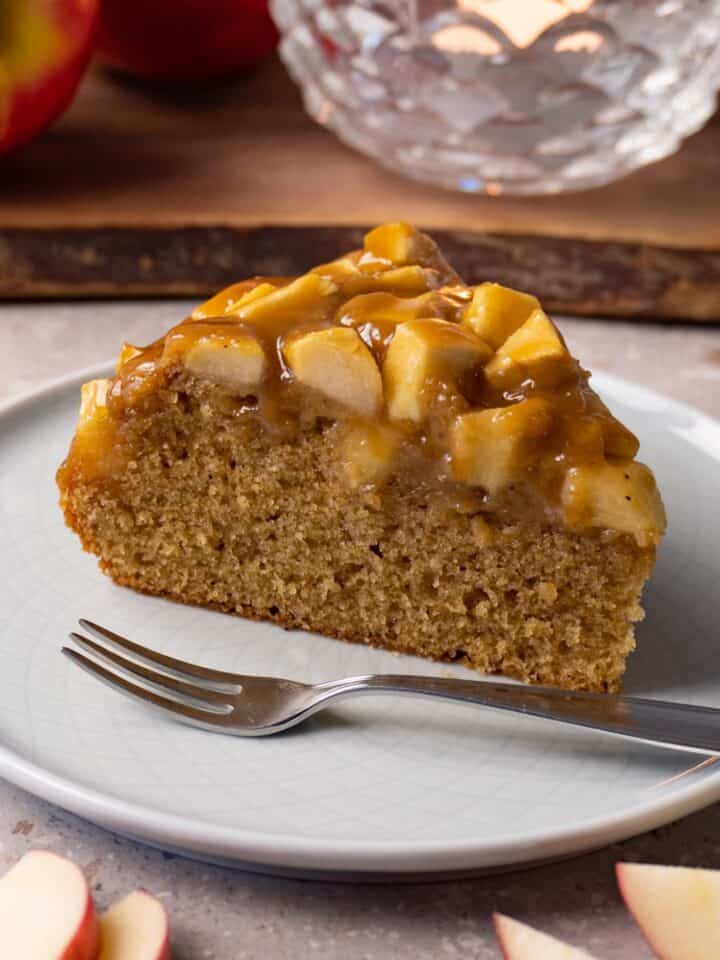A piece of apple cake topped with toffee sauce sits on a small round plate with a silver dessert fork.