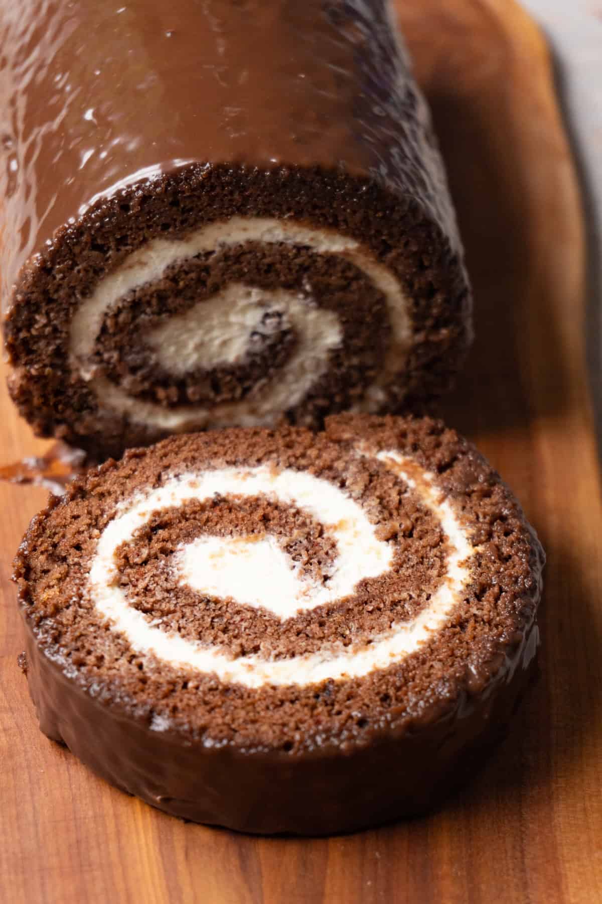 Chocolate Swiss roll cake with cream cheese filling is covered with dark chocolate glaze and served on a wooden serving board. One piece has been cut off and is lying on the same board.