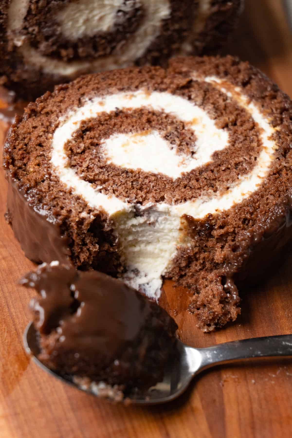 A piece of chocolate Swiss roll cake with cream cheese filling is covered with chocolate glaze and served on a wooden serving board. Several bites were taken from the piece.
